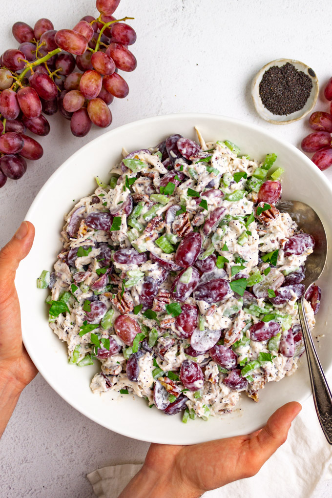 Sonoma chicken salad in a white bowl with hands serving hands holding the bowl and grapes off to the side.