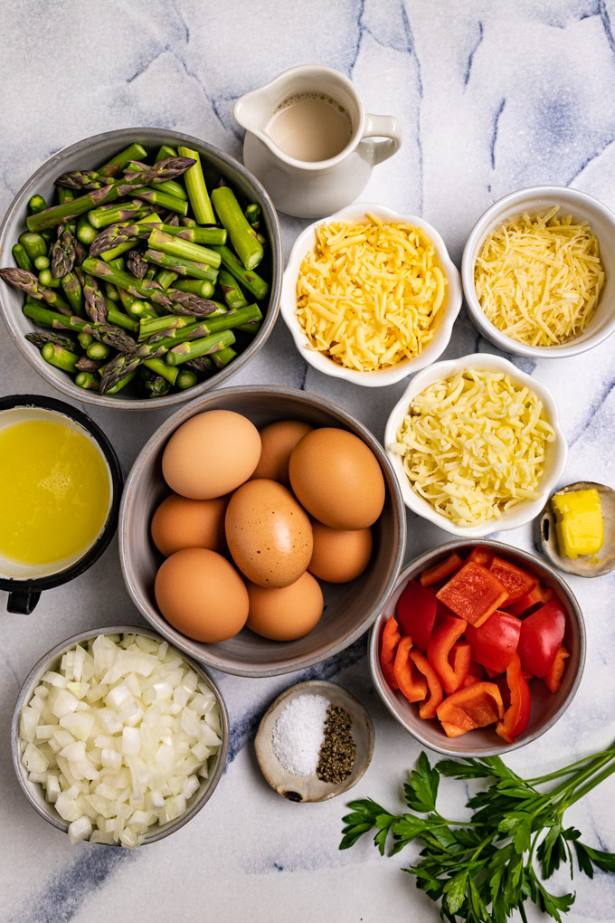 Ingredients for Asparagus frittata in bowls: asparagus, eggs, egg whites, cheeses, onions, red bell pepper, milk, butter, salt and pepper.