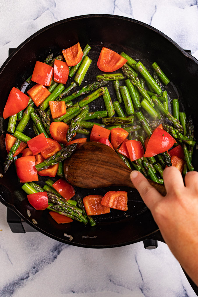 Asparagus and red bell pepper being sautéed in a cast iron skillet on a marble background. A hand is using a wooden spoon to stir the veggies.