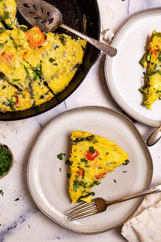 A slice of Asparagus frittata is on a gray plate with a fork beside it. Another plate is off to the side, only partially in the frame. The skillet of frittata is in left corner.