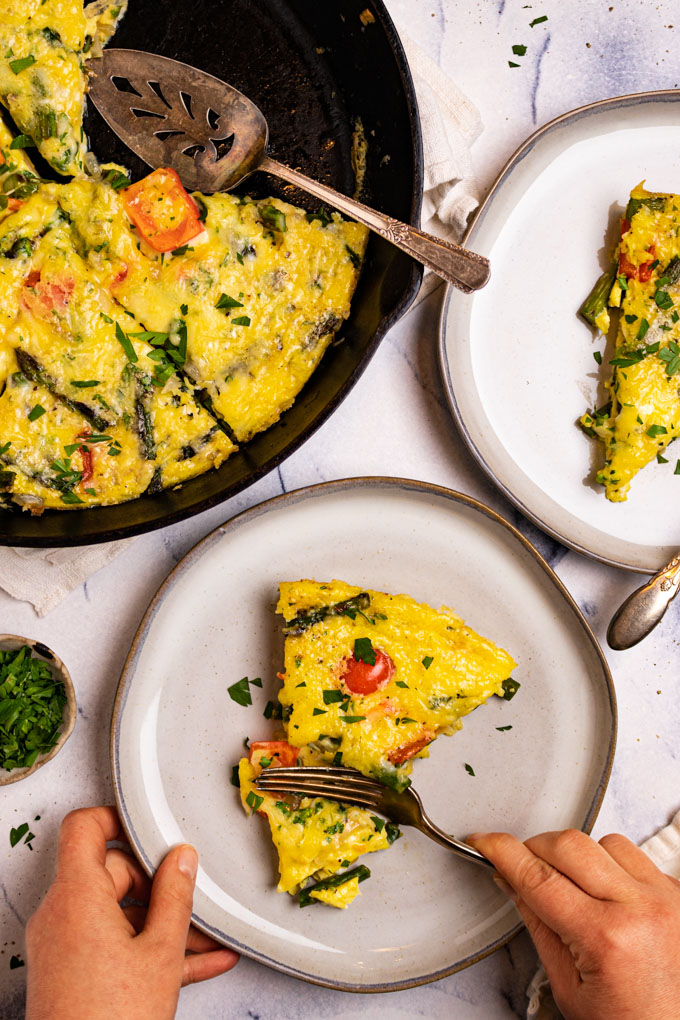 A slice of Asparagus frittata on a gray plate. A hand is holding the plate, another hand is cutting into the frittata slice with a fork. Another plate of frittata is cropped in the right corner. The pan of the remaining frittata is in the left corner.