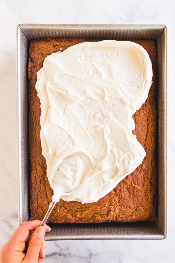 white frosting being spread onto a pumpkin sheet pan cake with a spoon