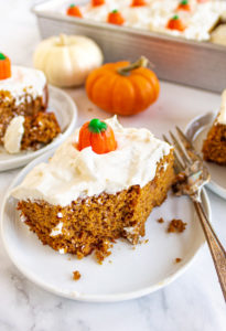 A slice of pumpkin cake, with white cream cheese frosting and a pumpkin candy on top, with a bite taken out of the cake.