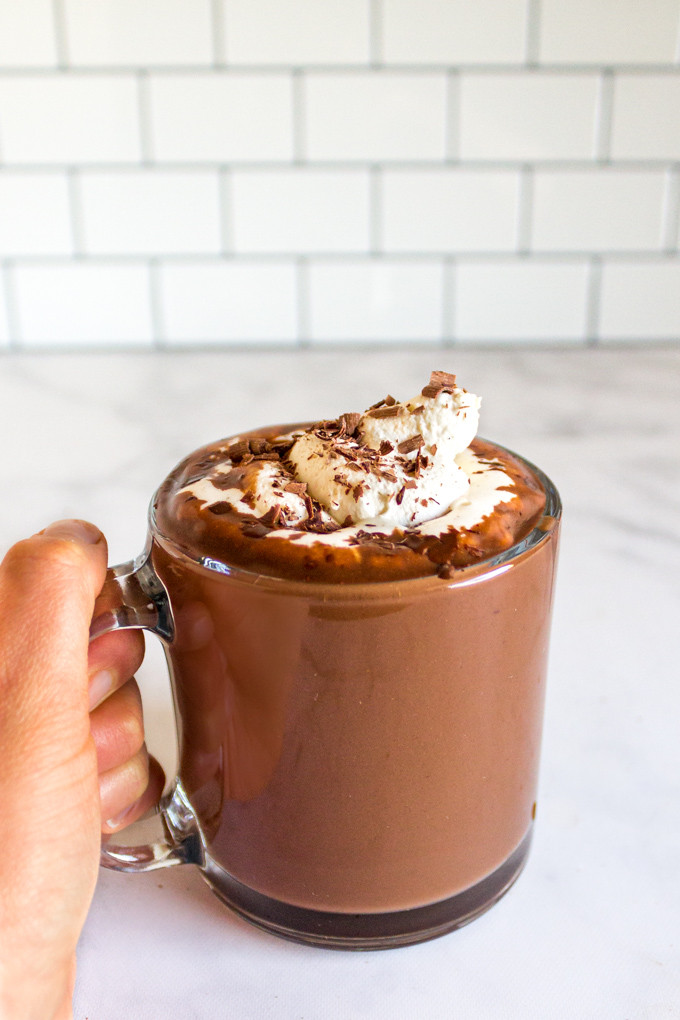 hot chocolate in a mug with partially melted whipped cream on top, and a hand holding the handle.