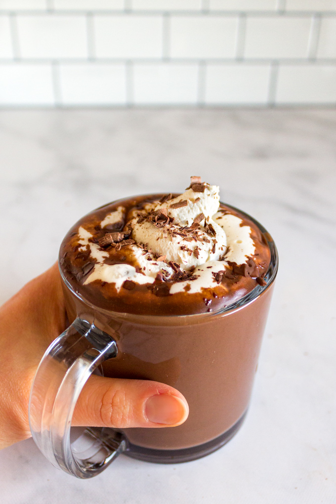 hot chocolate in a clear mug with a hand wrapped around the mug, with whipped cream on top.