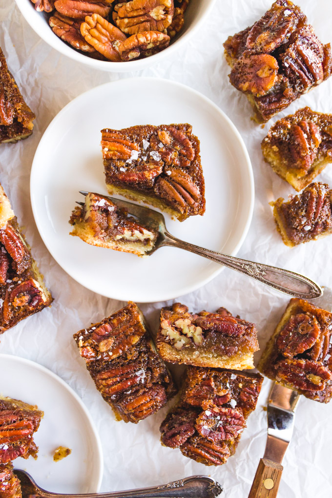 overhead of pecan pie bars on a white plate, with other slices of pecan pie bars around it, and a bowl of pecans in the backround.