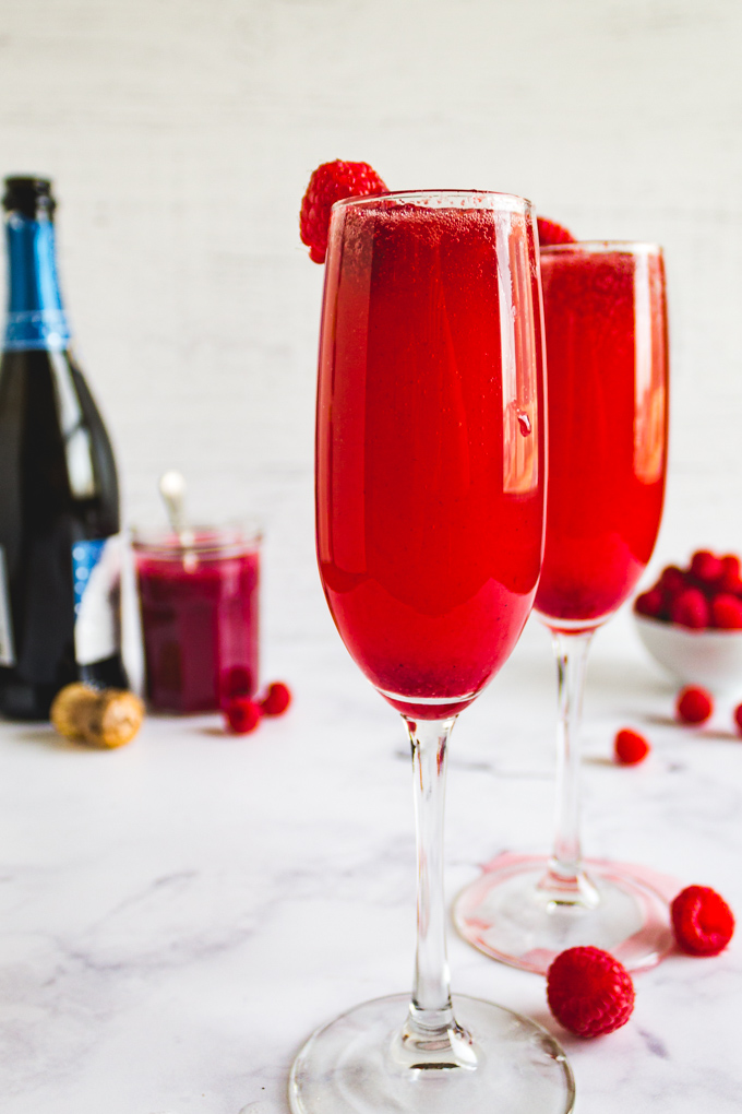 Raspberry bellini in 2 glasses, with a prosecco bottle, and raspberries in the background.