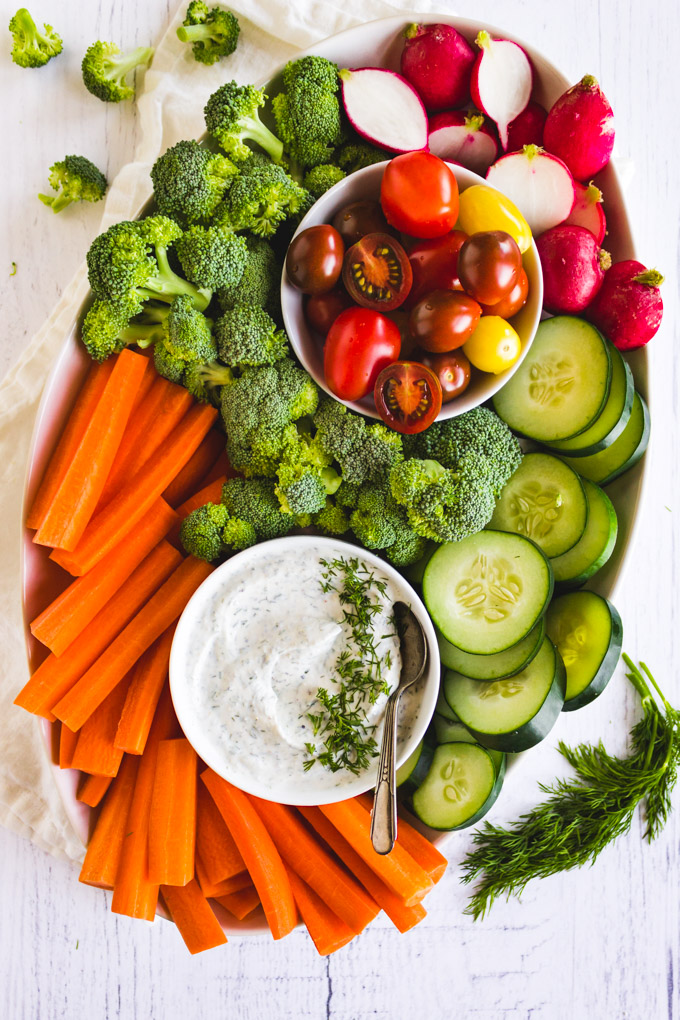 Platter of mixed raw veggies with a bowl of Greek yogurt dill dip and a spoon in it. On a white background.
