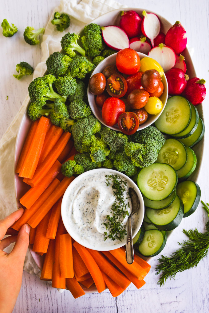 Raw veggies arranged on a large oval platter with a hand grabbing a carrot and a bowl of dill dip with a spoon.