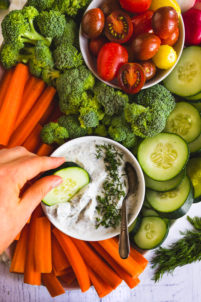 Close up of dill dip with a hand dipping a cucumber in it and raw veggies in the background.