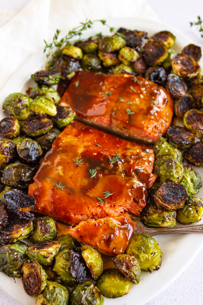 maple glazed salmon on a white oval platter with a fork taking a bite out of it and brussels sprouts around it.