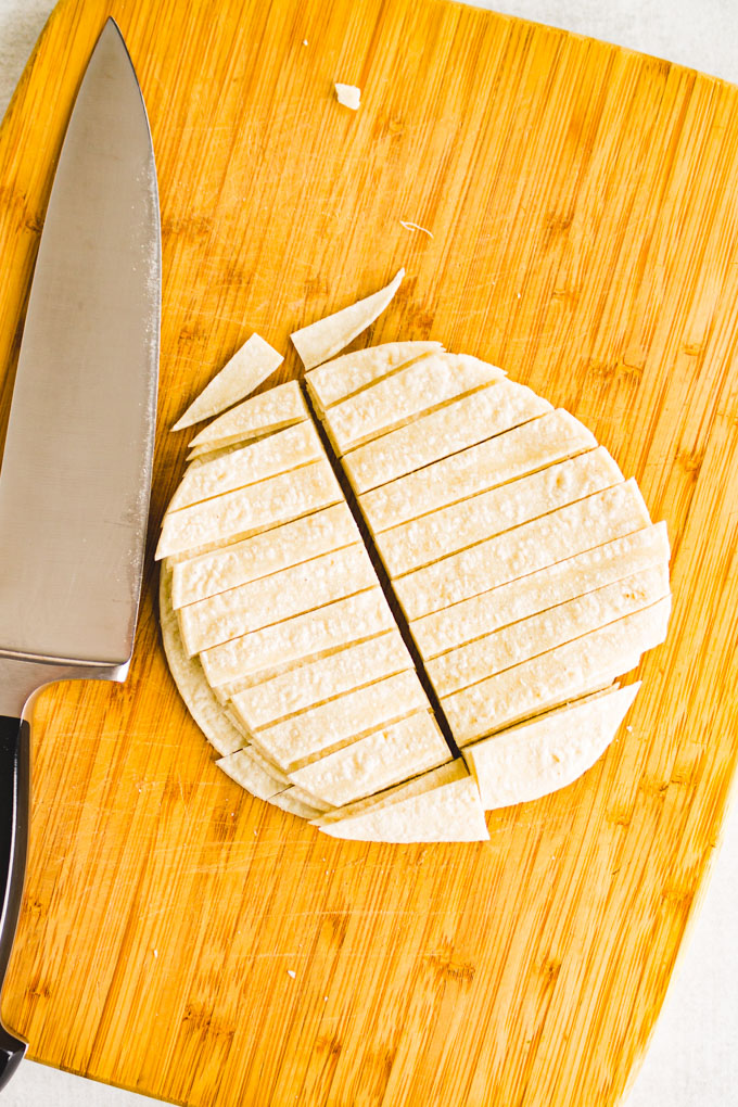 Corn tortillas sliced into strips, on a cutting board, with a chef's knife. 