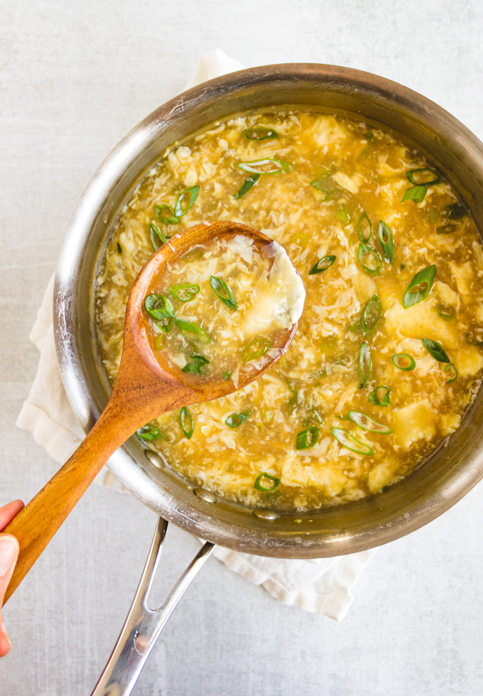 Egg drop soup on a light gray background in a sauce pan with a wooden spoon portioning some out.