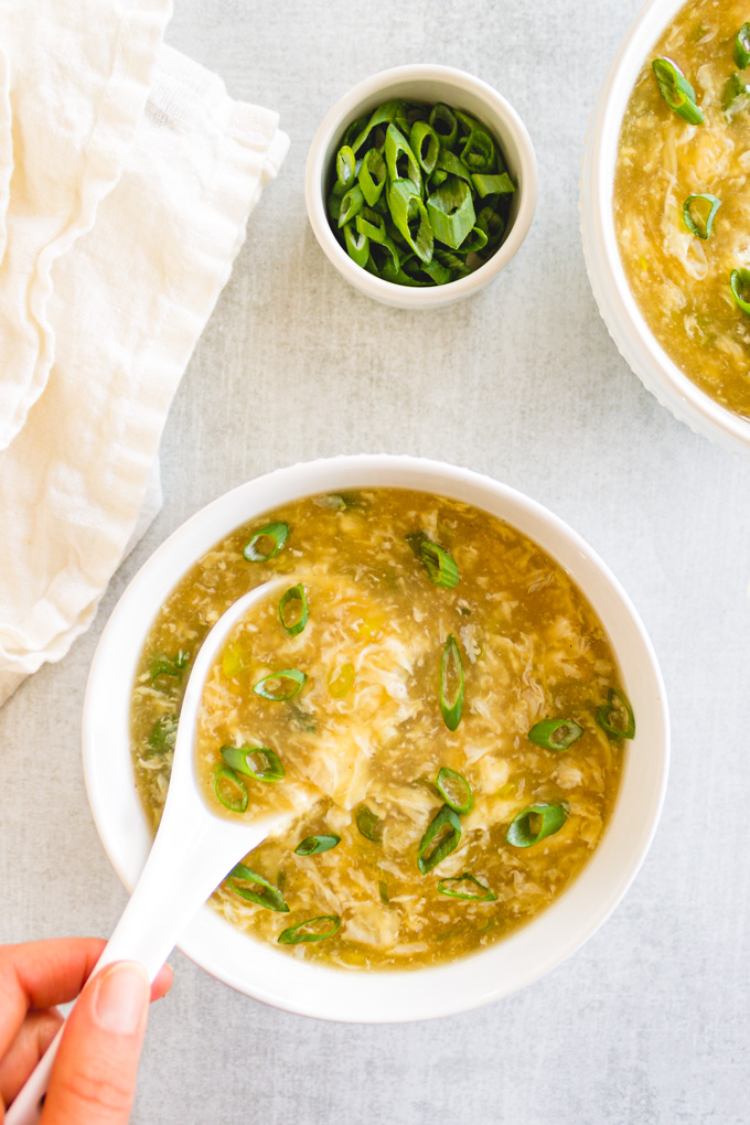 Egg drop soup in a white bowl, on a gray background with a spoon in the soup and a hand holding the soup.