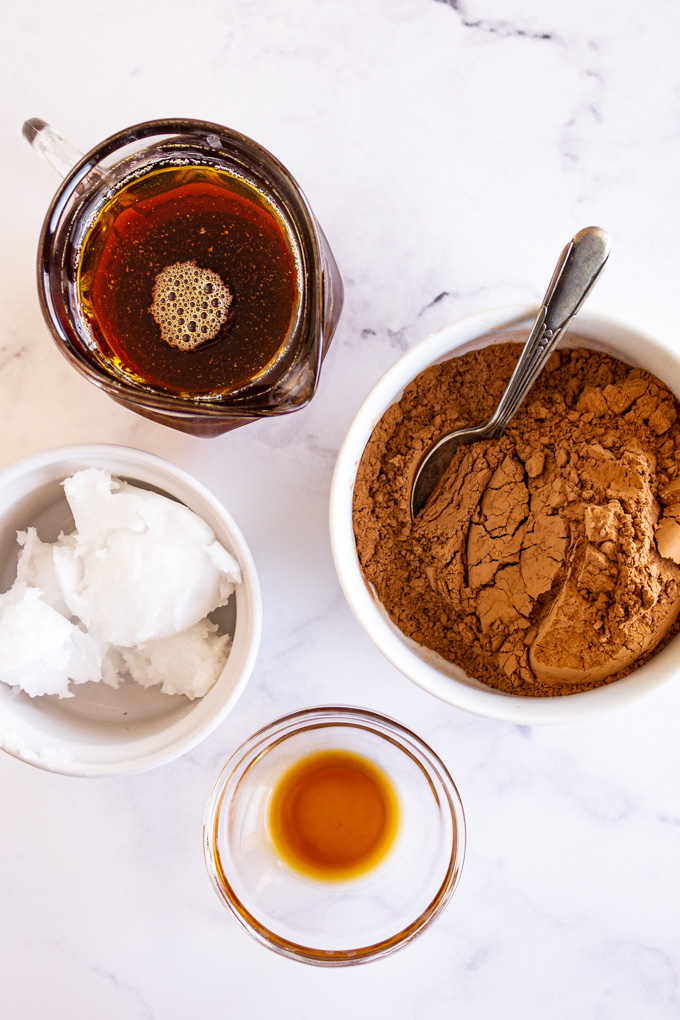 Cocoa powder, coconut oil, maple syrup, and vanilla in separate bowls on a white background.