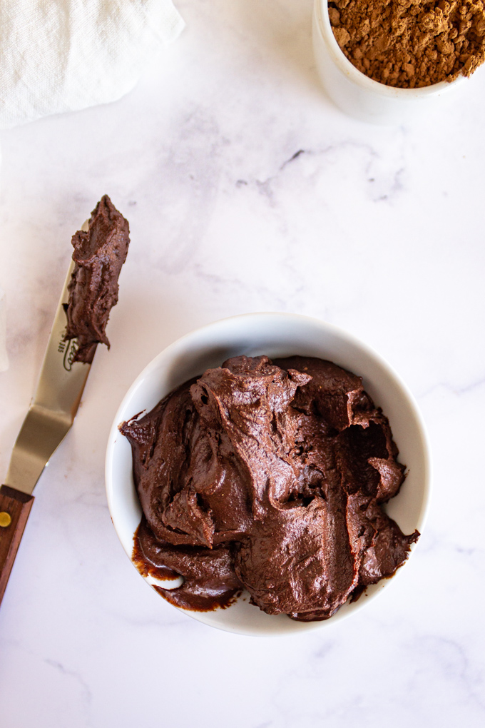 vegan chocolate frosting in a white bowl on a white background with some spread onto a spatula.