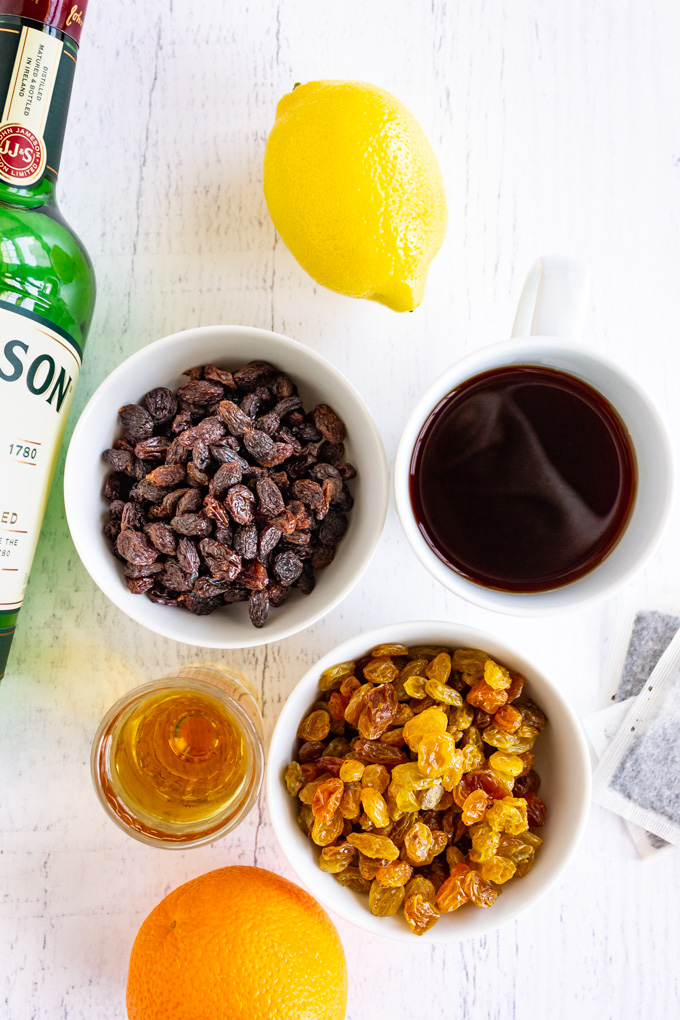 Raisins in white bowls, with a mug of tea, a shot glass of whiskey, a green Jameson bottle, and a lemon and orange on a white background.