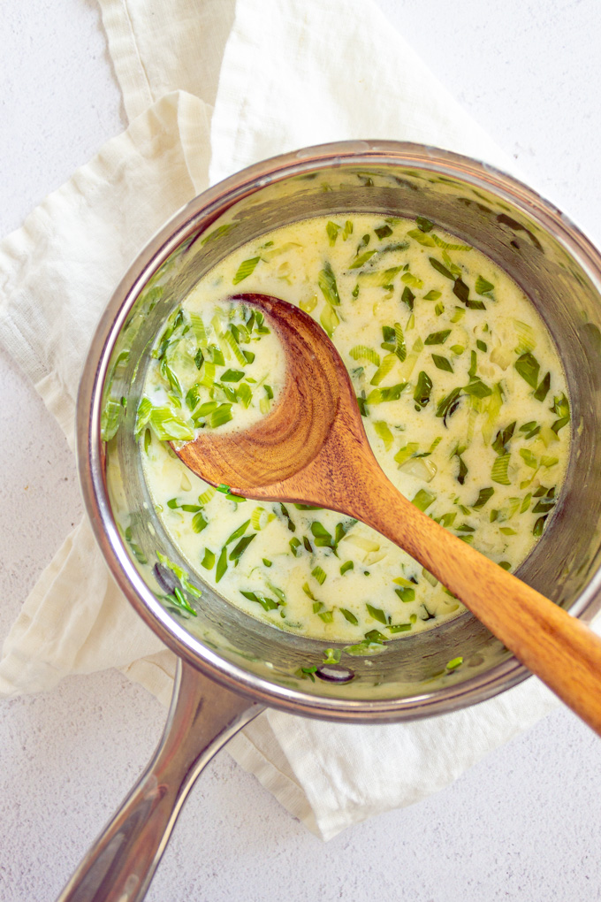 Milk in a small sauce pot with green onions, and a wooden spoon stirring it. On a white background.