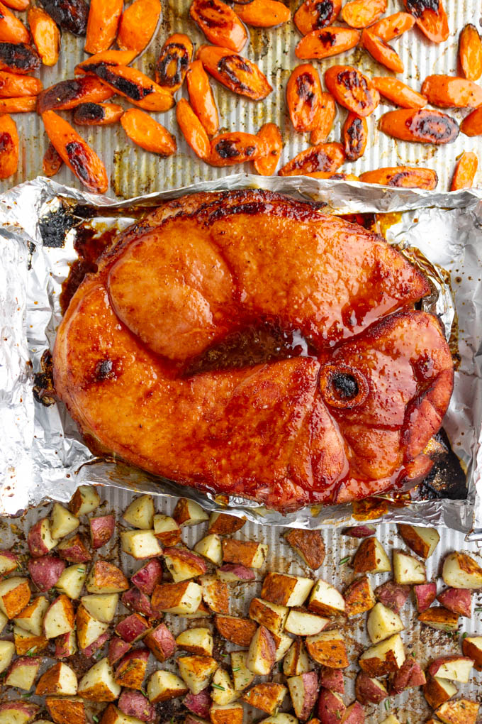 Roasted potatoes, roasted carrots, and a honey glazed ham steak on a sheet pan.