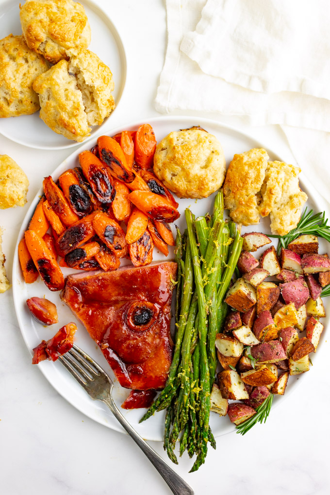 Sheet pan Easter dinner on a white plate. Ham with a fork taking a bite, honey butter carrots, buttermilk biscuits, roasted asparagus, and roasted potatoes. Extra buttermilk biscuits are in a plate in the background.