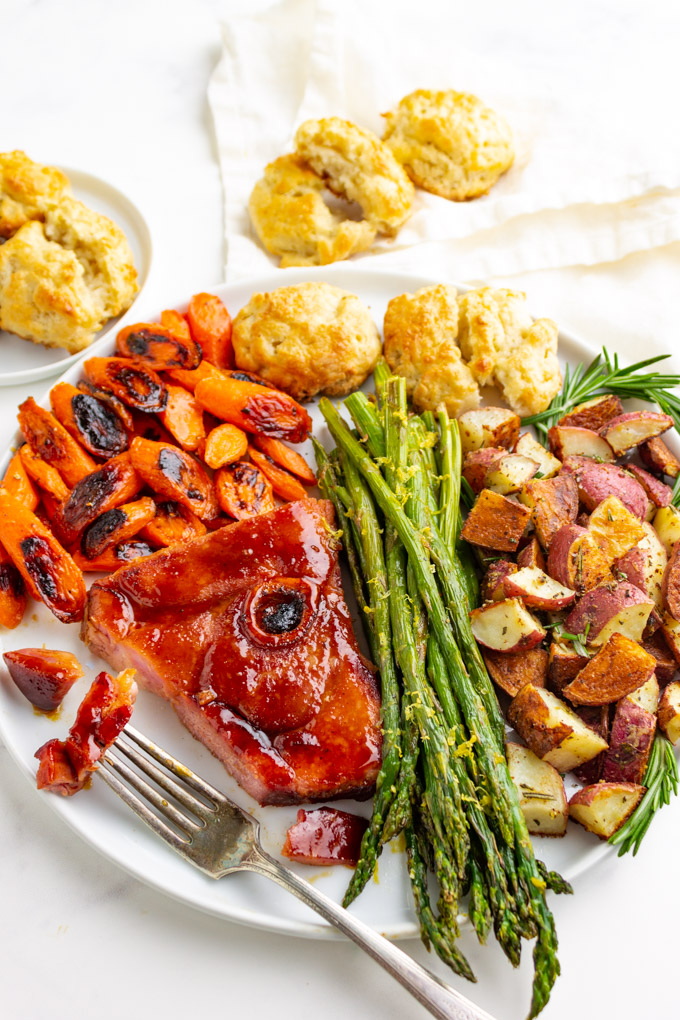 Sheet pan Easter dinner on a white plate. A slice of honey glazed ham, asparagus, roasted potatoes, honey butter carrots, and buttermilk biscuits.