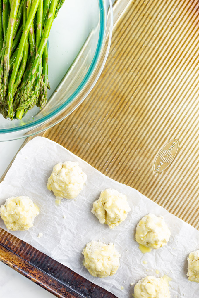 Buttermilk biscuits on a sheet pan with asparagus in a bowl.