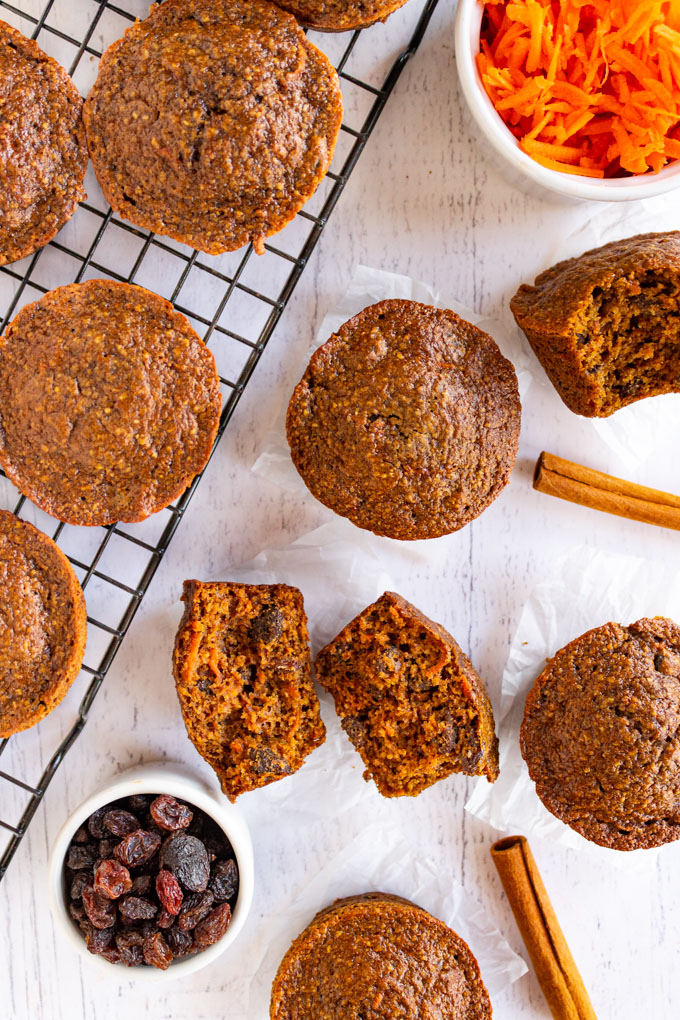 Healthy spiced carrot muffins on a white background. Some are on a wire cooling rack, some are split in half. There are raisins and shredded carrots in small bowls.