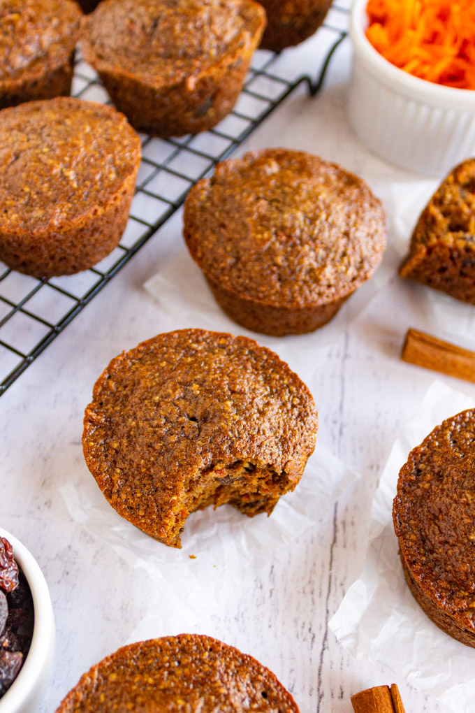 An angled shot of heathy spiced carrot muffins. One muffin has a bite taken out of it, some muffins are on a wire rack in the background.