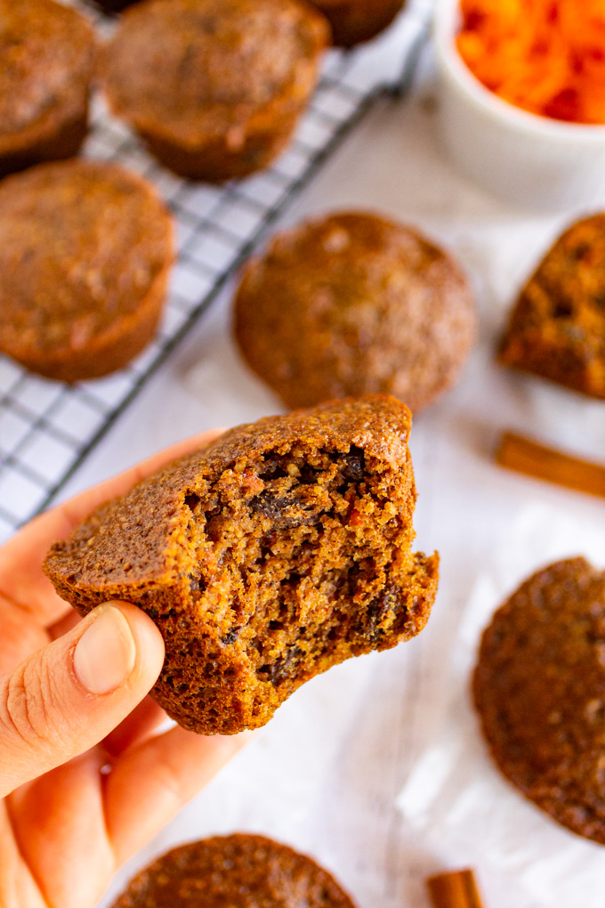 A close up of a hand holding a spiced carrot muffin with a bite taken out of it. There are more muffins blurred out in the background.