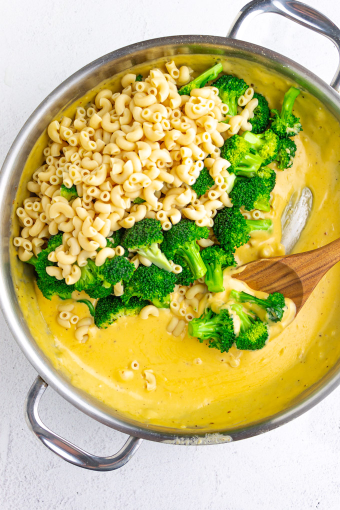 Yellow cheese sauce in a large stainless steel skillet. with pasta and broccoli being stirred in with a wooden spoon.