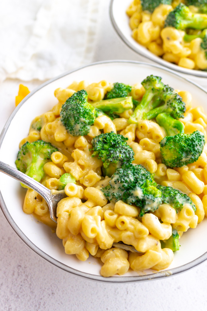 A close up angle shot of healthier mac and cheese with broccoli, in a white bowl, on a white background with a fork serving some mac and cheese up.