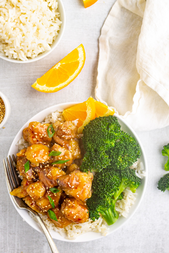 Lighter orange chicken in a bowl over white rice with steamed broccoli, on a gray background, with a bowl of white rice and orange sliced in the background.