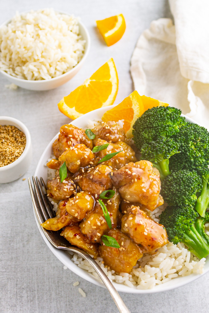 Lighter orange chicken in a white bowl, on top of rice, with steamed broccoli on the side.