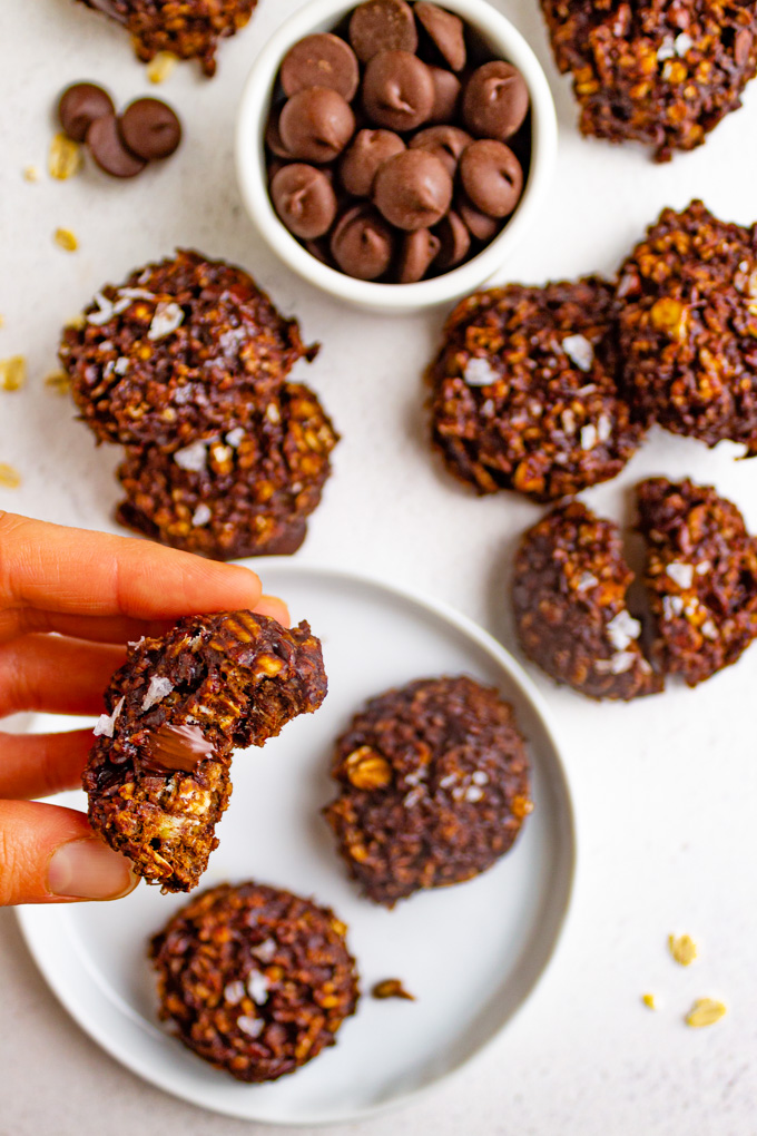 healthy chocolate oatmeal cookies on a white plate with a hand holding up one cookie with a bite taken out of it.