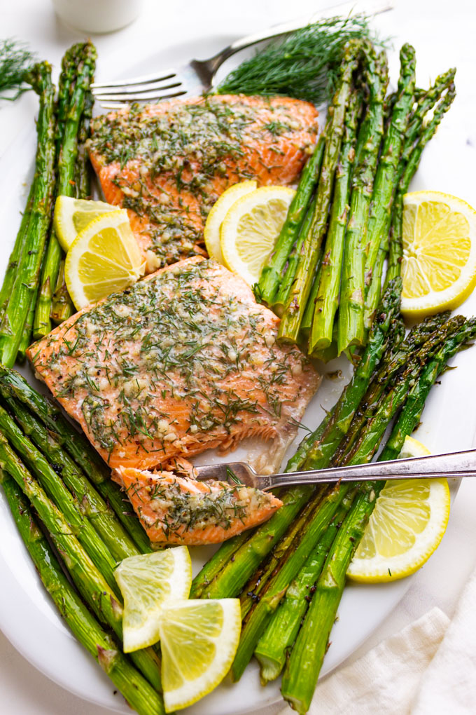 lemon dill salmon with asparagus on a white platter with a fork taking a bite out of the salmon