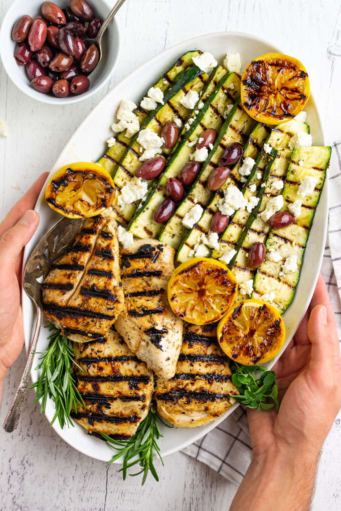 overhead shot of Greek grilled chicken with zucchini on a platter with hands holding it.