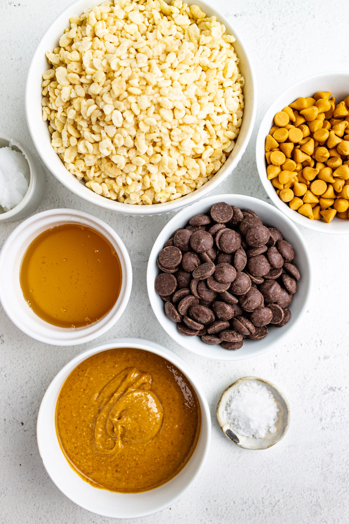 ingredients in separate white bowls - rice crispy cereal, butterscotch chips, chocolate chips, peanut butter, honey, and coconut oil.