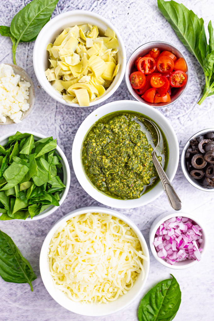 Ingredients in separate bowls. Pesto, shredded cheese, onions, olives, tomatoes, artichoke hearts, and spinach.