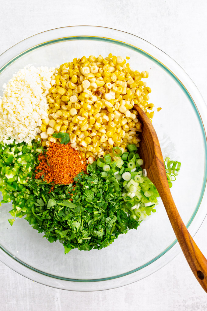 corn, diced jalapeno, chopped cilantro, green onion, and cotija cheese in a mixing bowl with a wooden spoon.