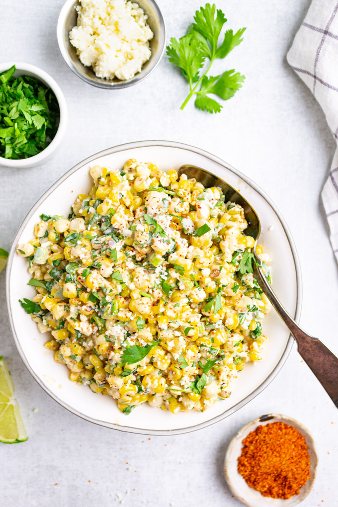 Mexican street corn in a white serving bowl with other ingredients in small bowls around it.