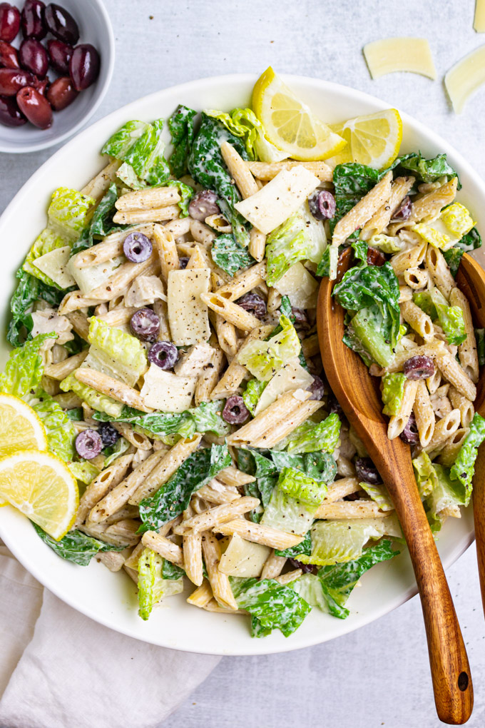 Caesar pasta salad in a large white serving bowl with wooden serving spoons.