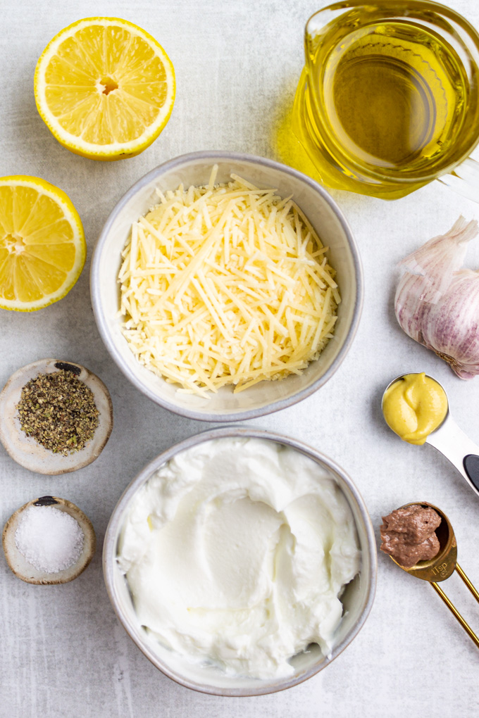 Ingredients in gray bowls - parmesan cheese, Greek yogurt, pepper, salt, dijon mustard, olive oil, lemons, garlic