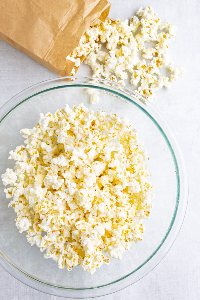 popcorn in a glass mixing bowl with popcorn spilling out of a brown paper bag in the background