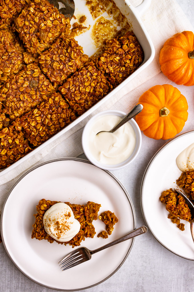 Squares of healthy baked pumpkin oatmeal on plates, with dollops of yogurt. There is a baking dish of pumpkin baked oatmeal in the background.