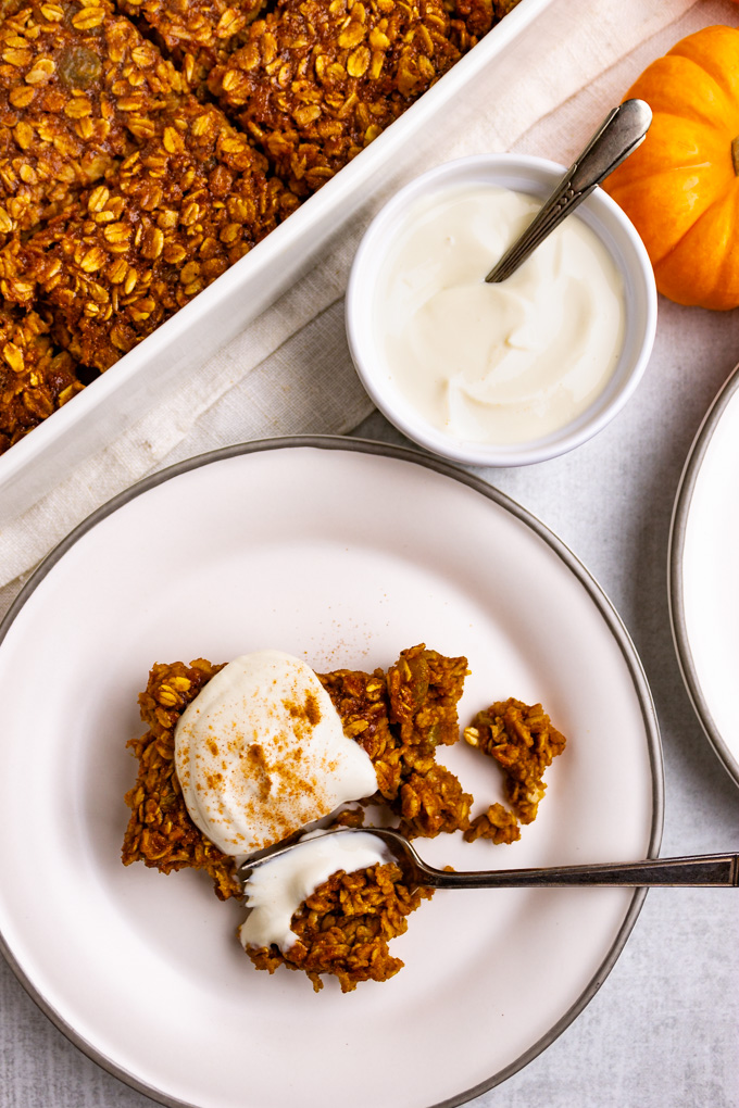 A slice of healthy baked pumpkin oatmeal on a plate with Greek yogurt topping on top, and a fork taking a bite out of it. A pan of baked oatmeal is in the background.