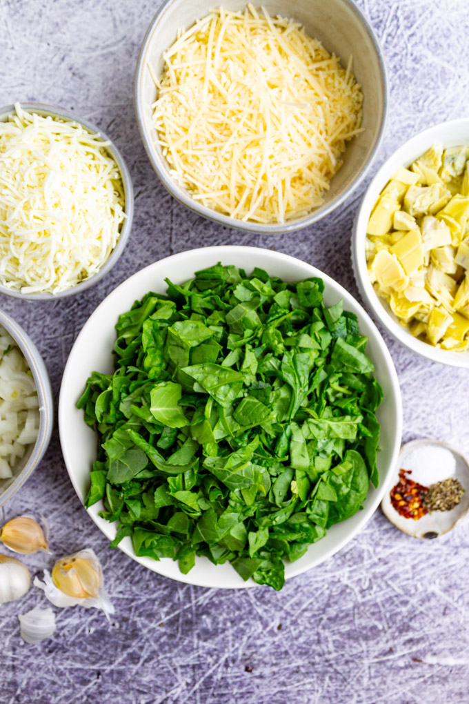 ingredients for spinach artichoke dip in bowls: spinach, spices, garlic, cheese, artichoke hearts, and onions