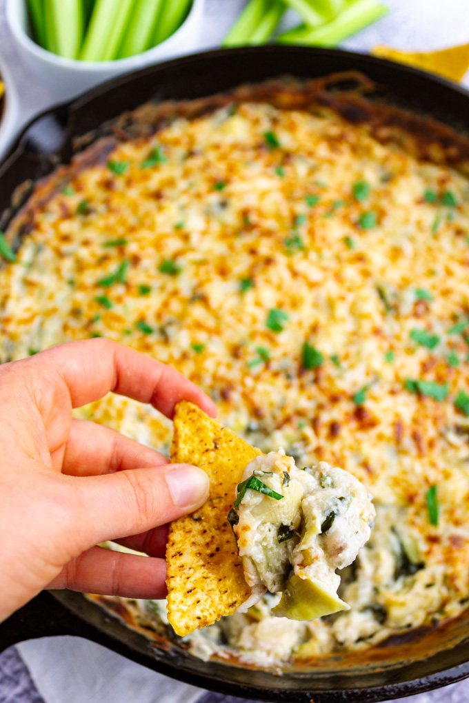 Closeup of a hand holding a chip with a big scoop of healthy spinach artichoke dip.