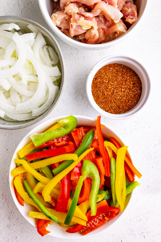 ingredients in bowls - sliced bell peppers, sliced onions, sliced chicken, fajita seasoning