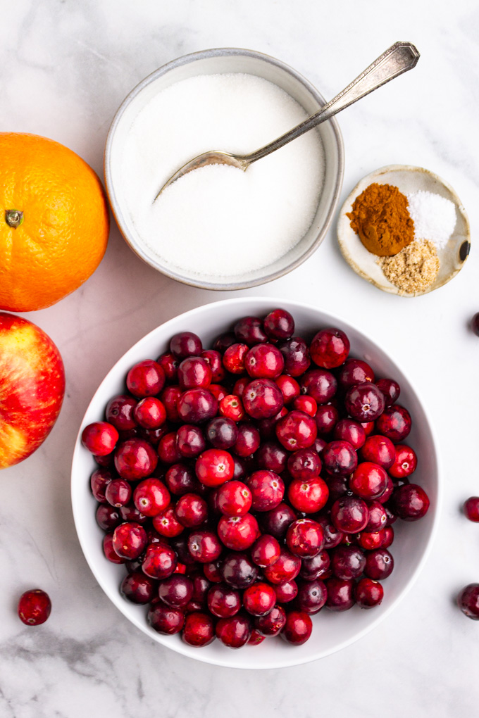 ingredients in separate bowls: whole cranberries, sugar. spices, an apple, and an orange.