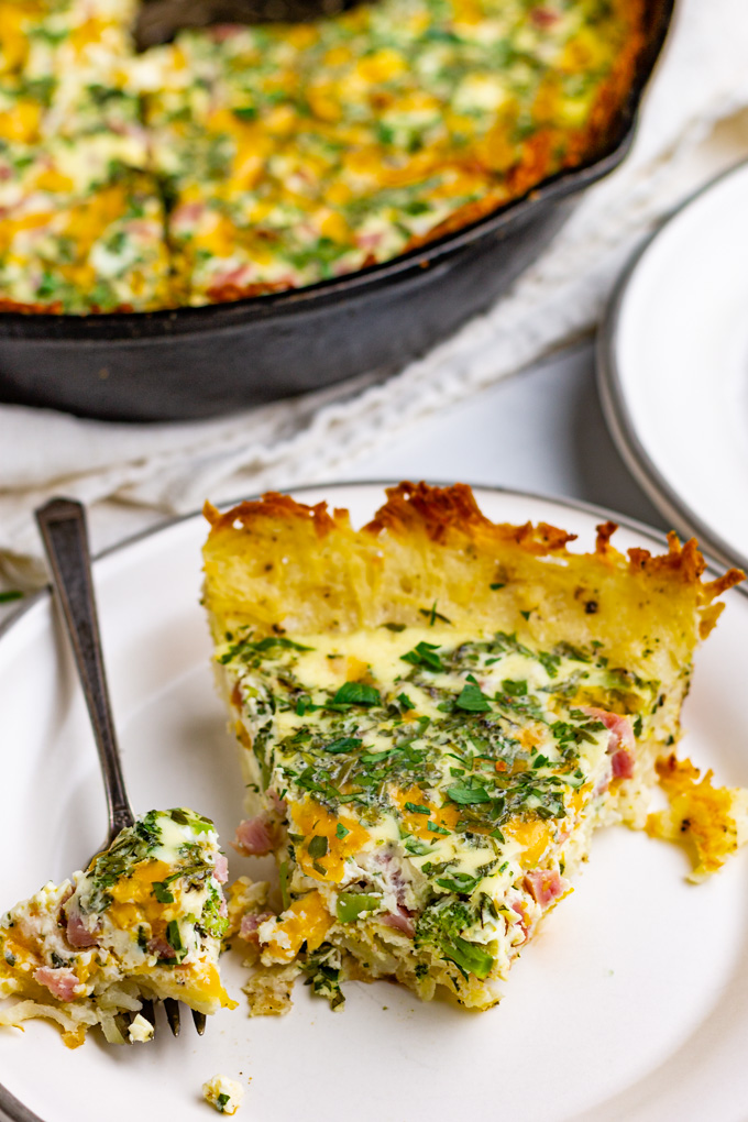 a close up shot of a slice of potato crust quiche with ham and broccoli with a bite taken out of the slice.